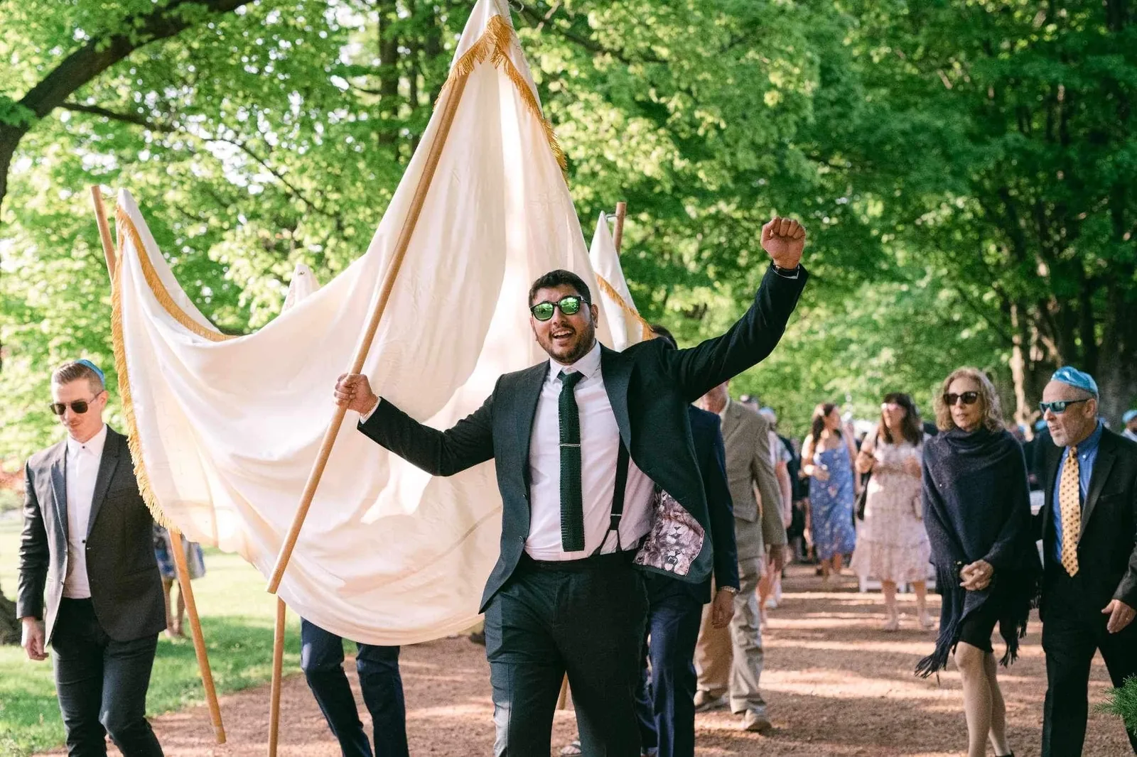 A chosson in a black suit walking to the chuppah with his arms raised, the canopy carried above him along a tree-lined path