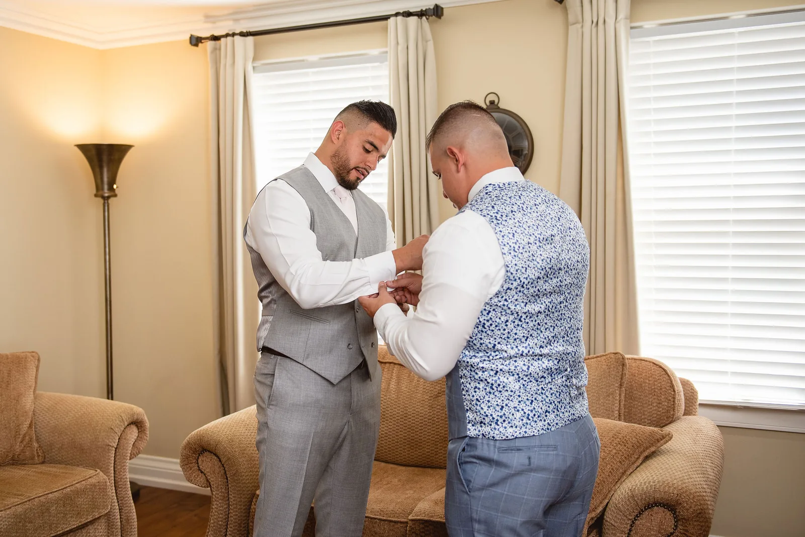 The groom in a grey waistcoat having his cuff fastened by a groomsman on the morning of the wedding