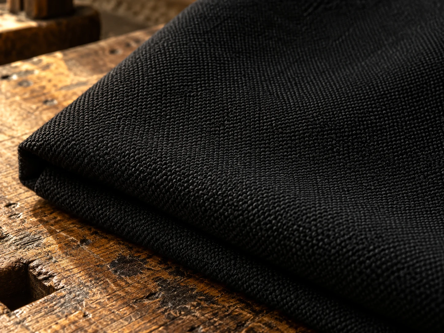 Macro photograph of black barathea dinner cloth showing its fine pebbled weave, folded on a worn oak bench in daylight