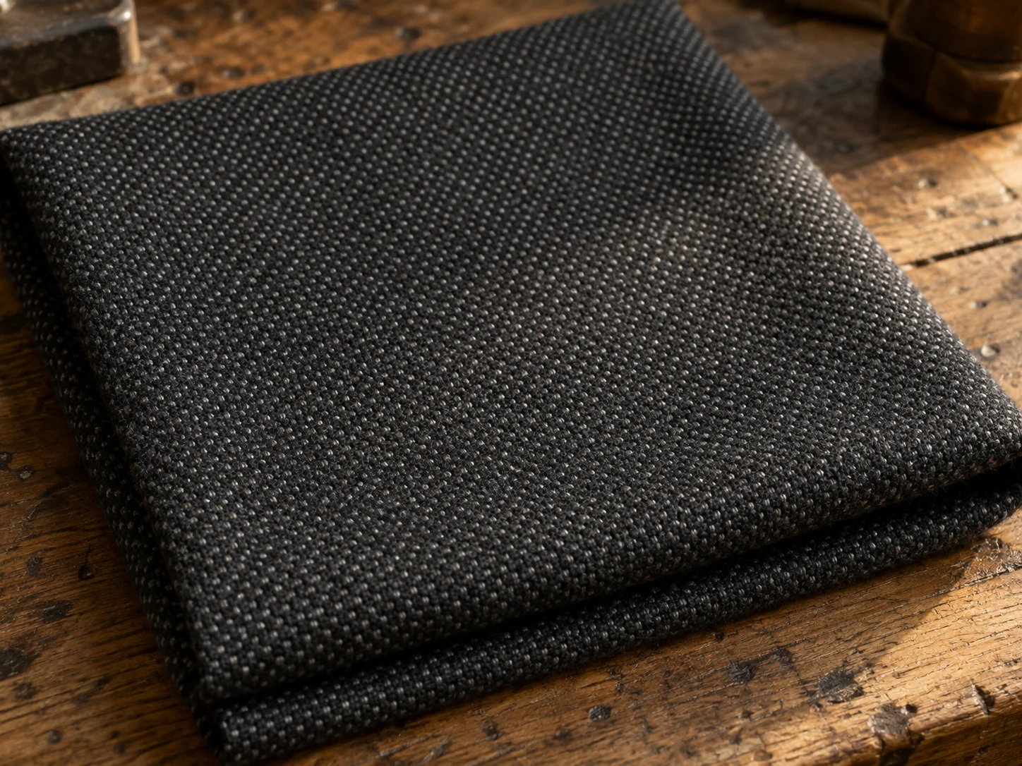 Macro photograph of charcoal worsted wool in a small all-over birdseye dot weave, folded on a worn oak bench in daylight