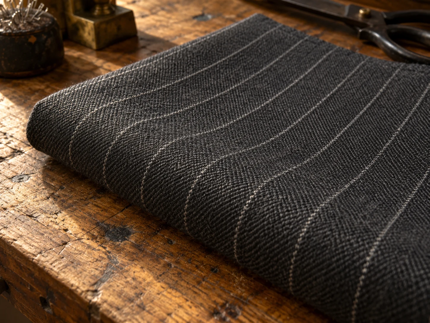 Macro photograph of charcoal worsted wool with a fine white pinstripe, folded on a worn oak bench in daylight