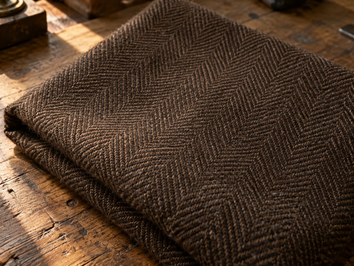 Macro photograph of chocolate brown wool in a herringbone zigzag weave, folded on a worn oak bench in daylight