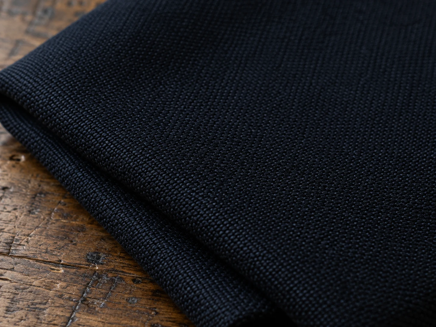 Macro photograph of midnight blue barathea dinner cloth showing its fine pebbled weave, folded on a worn oak bench in daylight