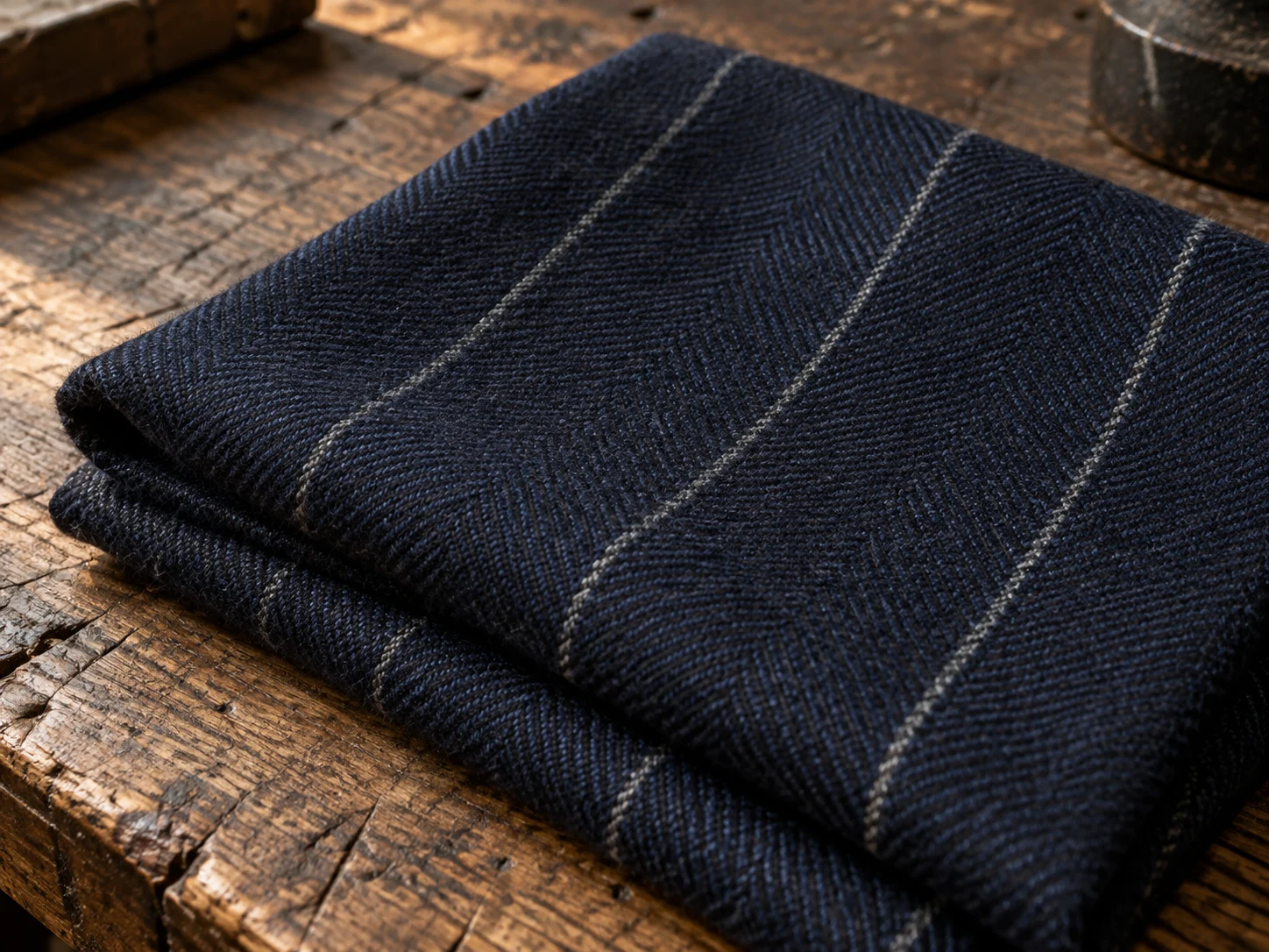 Macro photograph of navy worsted wool with a soft pale chalk stripe, folded on a worn oak bench in daylight