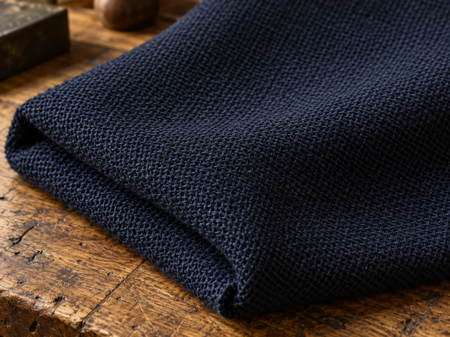 Macro photograph of navy high-twist fresco wool with a visibly open weave, folded on a worn oak bench in daylight
