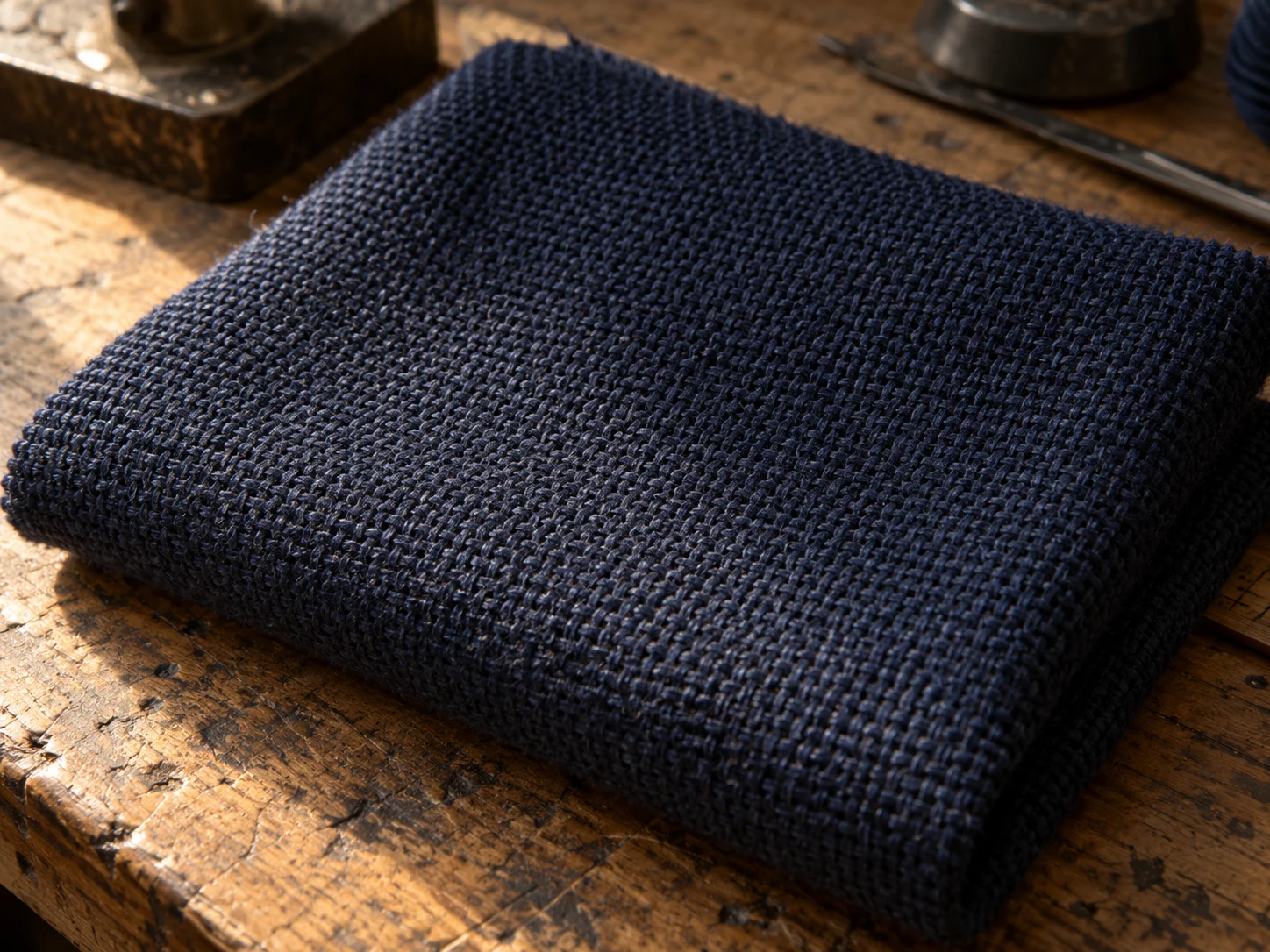 Macro photograph of navy wool hopsack in an open basket weave, folded on a worn oak bench in daylight