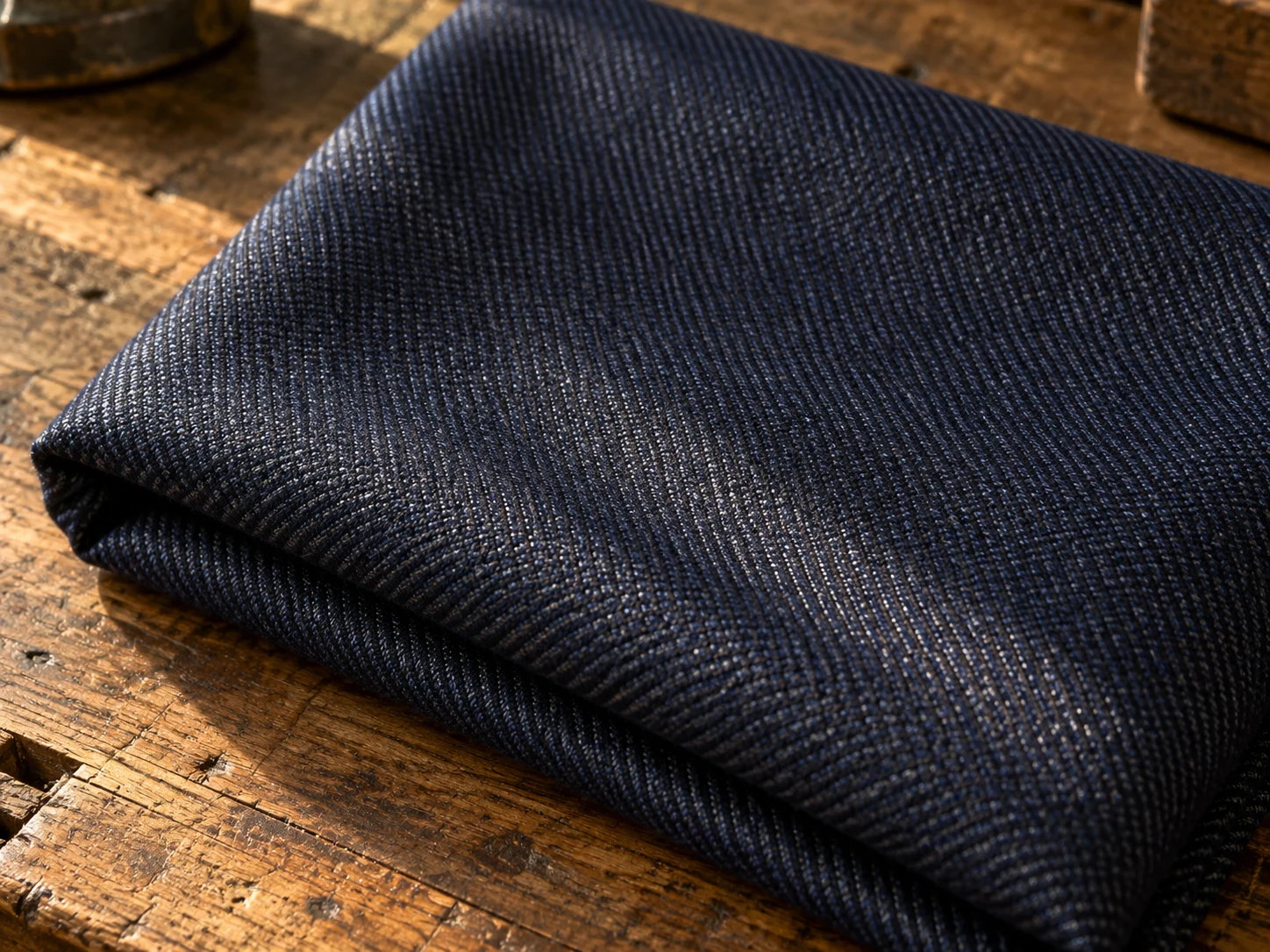 Macro photograph of navy worsted wool in a two-tone diagonal sharkskin weave, folded on a worn oak bench in daylight