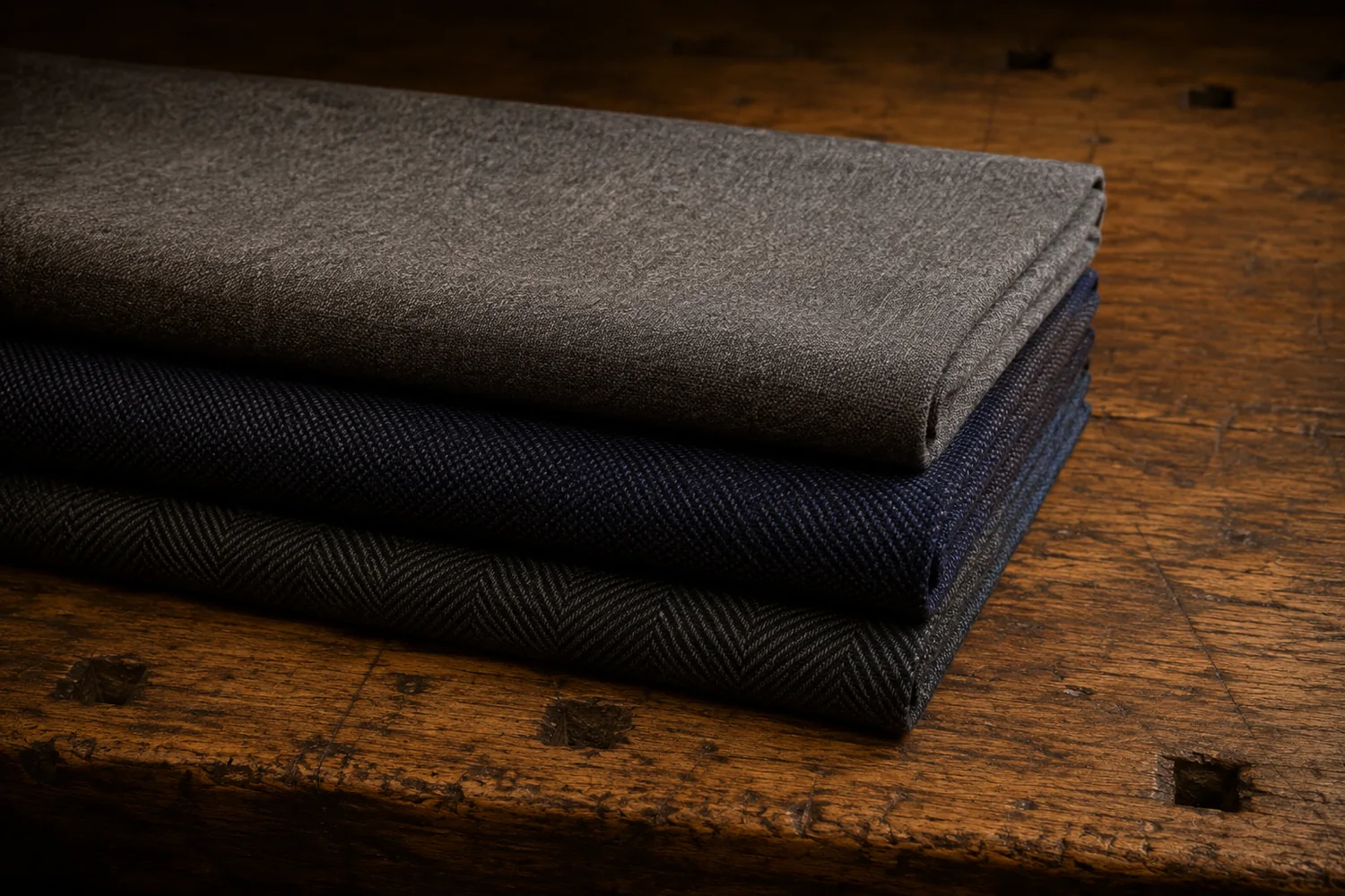 Three folded suiting fabrics stacked with their edges stepped: mid-grey, navy and a charcoal herringbone, each weave clearly different
