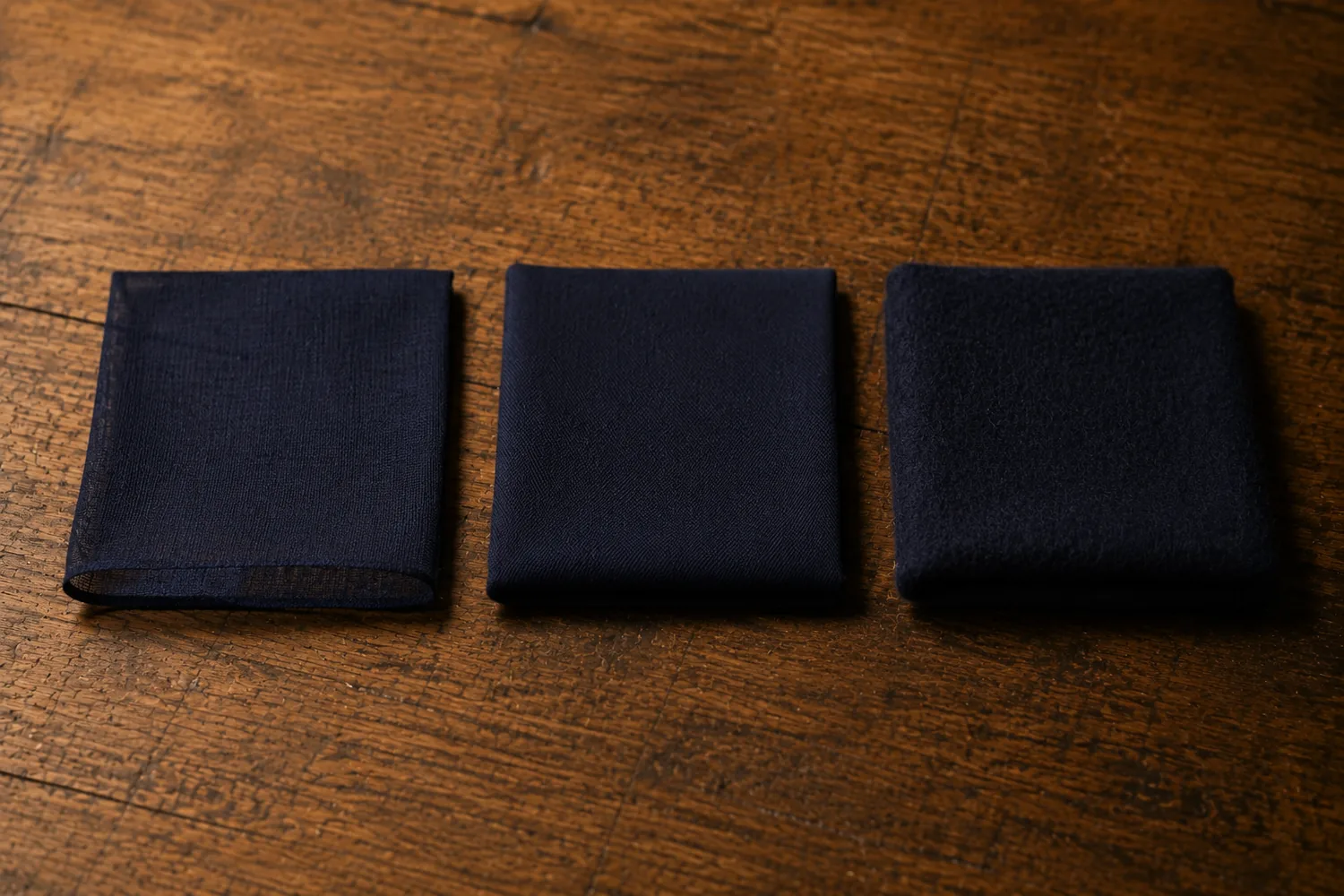 Three folded squares of navy suiting on an oak bench in order of weight: an open airy high-twist worsted, a smooth mid-weight worsted, and a dense brushed flannel