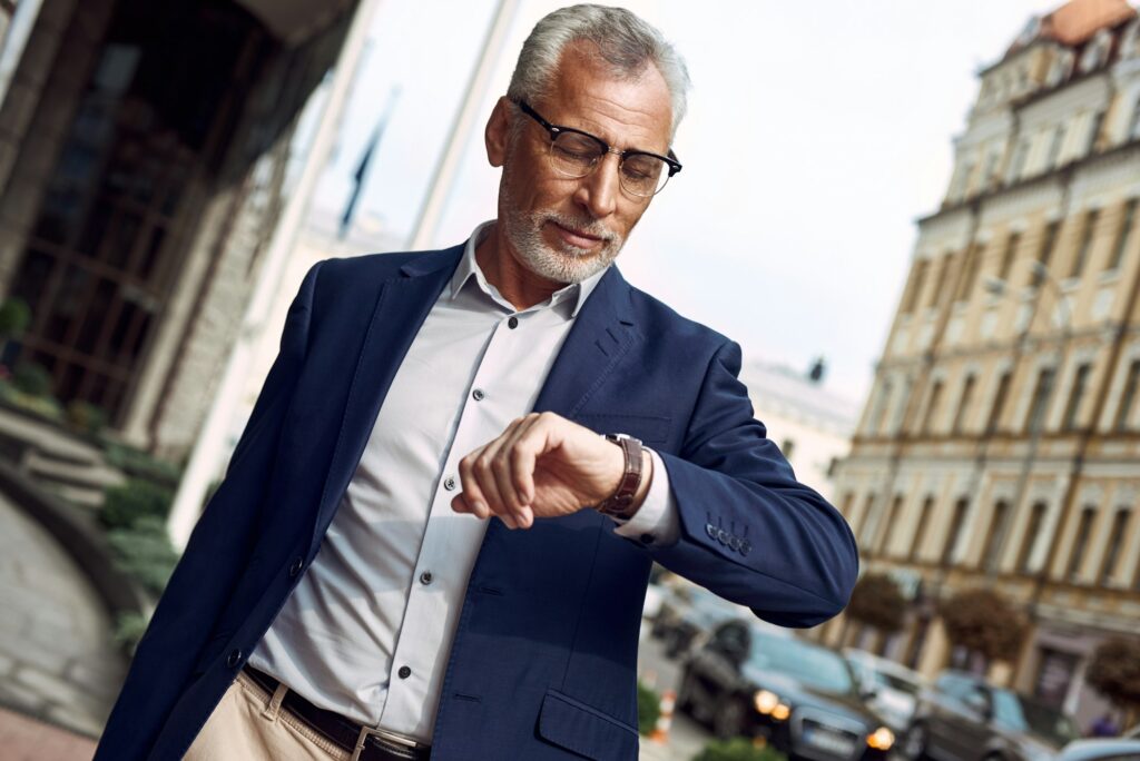 Thoughtful senior man in casual suit checking the time while standing outdoors